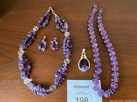 Overview of all pieces including amethyst chip bead necklace with clear beads, carved amethyst bead necklace, sterling silver amethyst pendant, and earrings on wooden surface