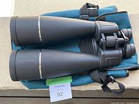 Black large binoculars resting on a blue carrying case, showing front view with large objective lenses and silver trim.