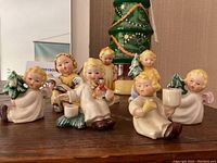 Photo showing front row of 7 collectible Christmas figurines including angels and children holding trees and musical instruments, arranged on wooden surface