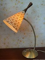 Full view of the mid century desk lamp showing the patterned conical shade, flexible neck, and metal base against a retro floral wallpaper background.
