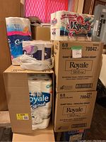 Photo showing stacked boxes of Royale Ultra Giant Roll toilet paper, a pack of Majesta toilet paper, and visible other packs in the background.
