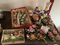 Wide view of the entire lot showing multiple ceramic china flowers arranged on a brown table surface, some boxed and some loose, including pink, red, yellow, and purple flowers with green leaves.