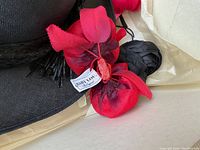 Close-up photo of a black woven straw hat with bright red and dark burgundy fabric flowers labeled 'Baby Lou' attached to the brim.