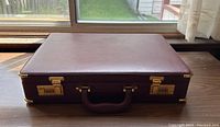 Top view of closed vintage burgundy leather briefcase with gold hardware on wooden table near window.