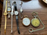 Overview of the lot showing two pocket watches on the right and five ladies wrist watches on the left with various strap and face styles arranged on a wooden surface.