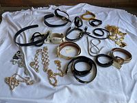 Full lot of 17 women's vintage belts shown on white cloth background, including leather straps and metallic chains arranged loosely to show variation in colors and styles.