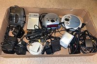 Overview of all audio devices and power adapters in a cardboard box, showing Sony Walkman, Caprice radio, Sylvania clock radio, Philips cassette player, Hipstreet digital player, cords and adapters.