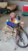 Photo showing multiple extension cords coiled inside a blue plastic storage bin, bundled ropes, and several bungie cords laid on the floor next to the bin.