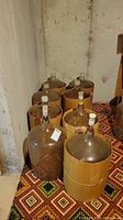 Eight large 24-inch diameter glass demijohn wine bottles, some wrapped in brown protective covers, arranged on a patterned carpet in a basement.
