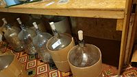 Photo showing eight glass demijohns lined up on the floor, some with cardboard protection around their bases and airlock stoppers fitted.