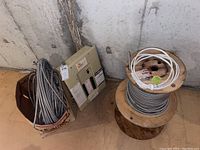 Overall view of cardboard box of conduit, two wooden spools (one loaded with conduit, one with white cable) and metal electrical box against concrete wall