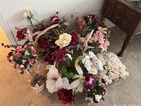 Full view of seven faux flower arrangements on carpet with various containers including pots, cups, and baskets. Flowers include roses and lilies in tones of pink, cream, white, and burgundy.