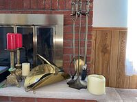Full view of fireplace tools, brass wood basket, candlesticks with red candles, and pillar candles arranged on hearth.