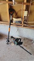 Photo showing Black and Decker electric hedge trimmer and Fiskars manual weed puller on concrete floor against wall.