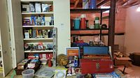 Photo of assorted hardware items including two metal toolboxes, spray cans, and glue tubes on wooden table with shelving in background.