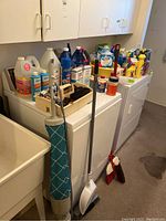 Photo showing cleaning solvent bottles, mop, broom, and dustpan arranged on top and beside a washing machine and utility sink.