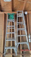 Two aluminum step ladders leaning against a wooden wall, one with a green Featherlite top tray and the other with a silver top tray.