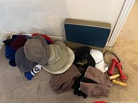 Photo showing assortment of men's hats, gloves, red hand weights, and a foam seat cushion arranged on floor.