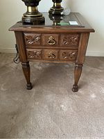 Photo of one wooden side table showing two drawers with carved fronts and metal hardware, glass top insert, and carved tapered legs.