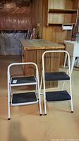 Front view of two folding utility step stools, white metal frames with black plastic steps shown side by side