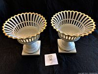 Pair of white porcelain pedestal baskets with open lattice sides and gold trim on scalloped edges seen from above on black background.