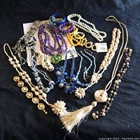 Full lot of 1970s colorful costume jewelry necklaces and bracelets laid on black background, showing variety of bead sizes, colors, and types including white, purple, blue, yellow, and brown beads, and several pendants and tassels.