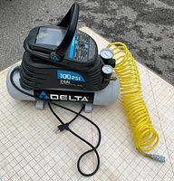 Delta CP200-1 air compressor viewed from side showing black and gray body, 2 gallon tank, 100 PSI capacity, and yellow coiled air hose with plug and fittings.