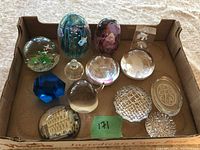 Full view of eleven assorted glass and acrylic paperweights in cardboard box showing diverse colors and patterns