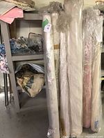 Five large bolts of vintage drapery fabrics wrapped in plastic standing upright on shelves in a storage space.