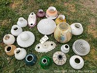 Photo showing 17 varied antique lamp shades of different colors, sizes, materials, and textures displayed on grass outdoors.