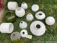 Twelve assorted antique ceramic and glass lamp shades in white and green, placed on grass.