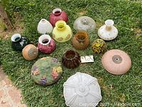Photo showing a group of thirteen different antique lamp shades arranged on grass, showcasing a variety of colors, textures, shapes and sizes.