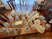 Photo of approximately 38 various styles of white glass lamp shades laid out on a wooden table showing diverse sizes and decorative patterns.