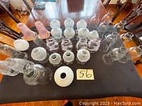 Top-down view of 24 glass lamp shades in various styles, including clear, frosted, pink tinted, textured, and patterned glass arranged on a black rectangular cloth on a wooden table.