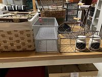Collection of assorted baskets and bins displayed on a table showing woven, metal mesh, and wire construction.