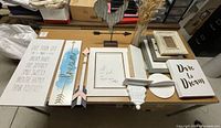 View showing home decor collection including signs with inspirational sayings, picture frames, wooden arrows, metal rooster figure, decorative corbel, glass vase with dried grasses, and small shelves.