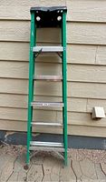 Green metal step ladder with black top tray leaning against beige siding wall. Four steps visible.