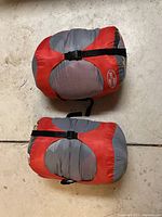 Photo of two compact sleeping bags in red and gray secured with compression straps on concrete floor.