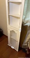 Photo of white wooden corner shelf showing three triangular open shelves above one lower cabinet with mesh panel door, placed beside a curtain and wall.