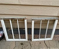 Two small vintage white wooden window frames each with four vertical glass panes, placed on ground leaning against siding.