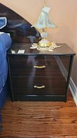Front view of first laminate wood veneer bedside table with two drawers and gold-tone handles, with a floral glass shade lamp sitting on top beside a bed.