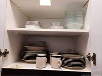 Shelf with stacked white plates, black and white plates, coffee mugs, and clear glass bowls