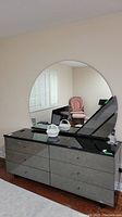 Side view of retro six-drawer dresser with laminate finish and large round mirror mounted behind it. Shows light reflection and room interior.