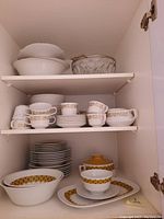 Cupboards showing neatly stacked white porcelain plates and bowls with mustard yellow and gold geometric bands, stacked teacups with the same pattern, also white serving plates and mixing bowls