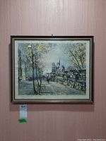 Framed print depicting a city street scene with trees and a cathedral in the background, hung on a pink textured wall.