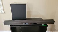 Black rectangular Sony soundbar with control panel and black subwoofer behind against a wall on a black surface.