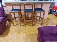 Front view of three wood frame stools with black upholstered seats arranged side by side in front of kitchen counter.