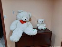 Two white teddy bears sitting on wooden cabinet, one large with red bowtie and one smaller.