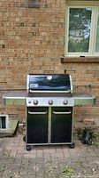 Front view of Weber Genesis natural gas BBQ grill showing three control knobs, stainless steel lid with thermometer, side shelves, and lower storage on wheels.