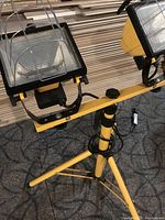 Photo of two yellow construction flood lights with black frames mounted on an adjustable yellow tripod stand, showing front and top angle.